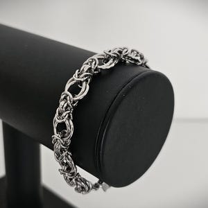 May include: A silver-toned bracelet with a woven chain design, displayed on a black jewelry stand. The bracelet features a series of interconnected links, creating a textured and intricate pattern. The background is a soft, neutral color, highlighting the bracelet's details.