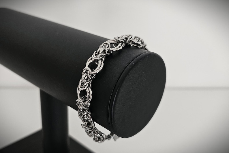 May include: A silver-toned bracelet with a woven chain design, displayed on a black jewelry stand. The bracelet features a series of interconnected links, creating a textured and intricate pattern. The background is a soft, neutral color, highlighting the bracelet's details.