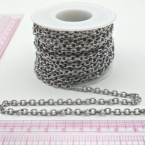 Może przedstawiać: Szpula srebrnego łańcucha z rozłożonym odcinkiem. Łańcuch składa się z małych, okrągłych ogniw. Różowa linijka znajduje się w tle. Łańcuch jest prawdopodobnie używany do tworzenia biżuterii lub rękodzieła.