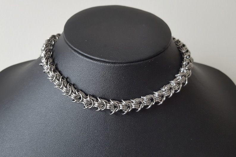 Pode incluir: Um colar de malha de corrente prateado, exibido em um busto de manequim preto. O colar &eacute; feito de pequenos an&eacute;is de metal interligados, criando uma apar&ecirc;ncia texturizada e tecida. O design de malha de corrente forma um gargantilha.