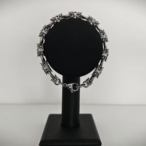 May include: A silver-colored bracelet with an intricate chain design is displayed on a black stand. The bracelet features a series of interconnected links, creating a textured appearance. The clasp is visible, and the background is a soft, neutral tone.