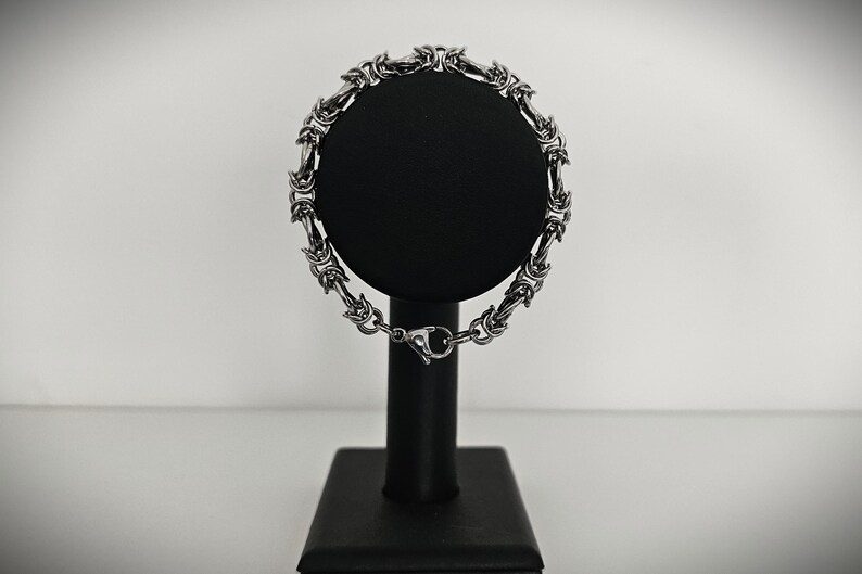 May include: A silver-colored bracelet with an intricate chain design is displayed on a black stand. The bracelet features a series of interconnected links, creating a textured appearance. The clasp is visible, and the background is a soft, neutral tone.