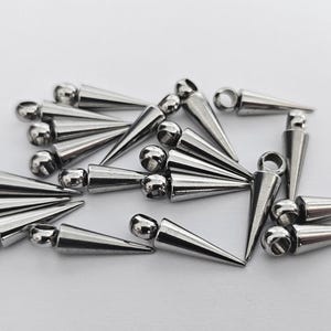 May include: A collection of silver-colored metal cone-shaped spikes with a rounded top and a small loop at the base. The spikes are scattered on a white surface, reflecting light.