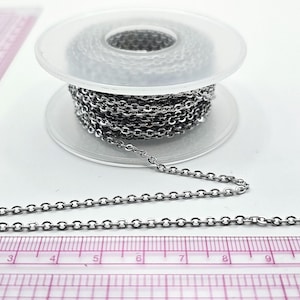 May include: A spool of silver-colored chain with a small oval link design. The chain is partially unraveled, lying next to a pink ruler with inch markings. The chain is likely used for jewelry making or crafts.