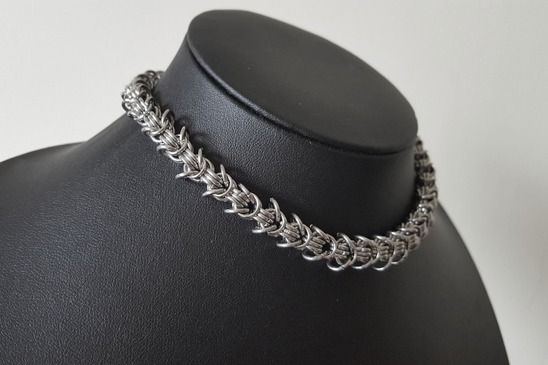 Pode incluir: Um colar de cota de malha prateado exibido em um manequim preto. O colar &eacute; feito de pequenos an&eacute;is de metal interligados, criando uma apar&ecirc;ncia texturizada e tecida. O design &eacute; um estilo gargantilha, perto do pesco&ccedil;o.