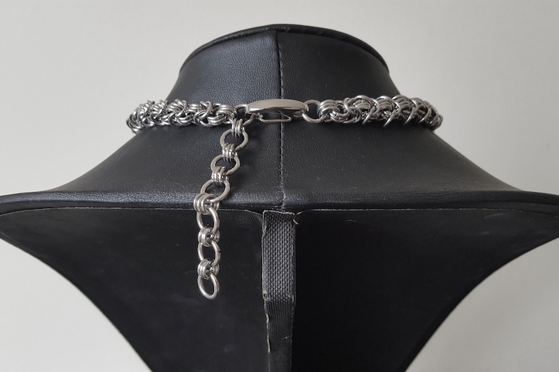 Pode incluir: Um colar prateado com um design de malha de corrente, exibido em um manequim preto. O colar apresenta um fecho de lagosta e uma corrente ajust&aacute;vel. A joia &eacute; feita de metal e &eacute; adequada para v&aacute;rias ocasi&otilde;es.