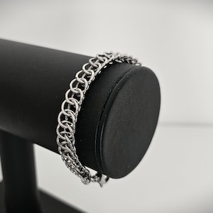 Puede incluir: Una pulsera de cota de malla plateada exhibida en un expositor de joyas negro. La pulsera está hecha de anillos de metal entrelazados, creando una apariencia texturizada y tejida. El fondo es blanco suave y borroso.