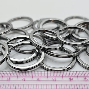 Puede incluir: Un conjunto de llaveros divididos de color plateado, algunos abiertos y otros cerrados, se encuentran dispersos sobre una superficie blanca. Una regla rosa y blanca está en primer plano. Los llaveros son de metal y tienen un diseño dividido.