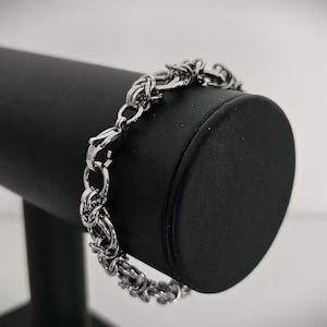 May include: A silver-colored bracelet with a chain-link design is displayed on a black jewelry stand. The bracelet features a series of interconnected links, creating a textured appearance. The clasp is visible, and the bracelet is presented against a blurred background.