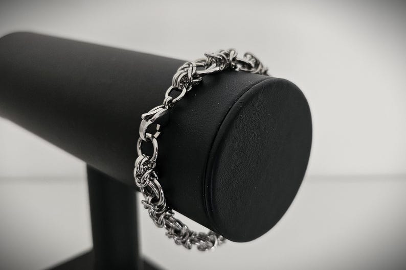May include: A silver-colored bracelet with a chain-link design is displayed on a black jewelry stand. The bracelet features a series of interconnected links, creating a textured appearance. The clasp is visible, and the bracelet is presented against a blurred background.