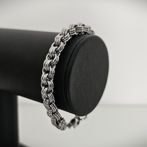 May include: A silver-colored bracelet with a thick, textured chain design. The bracelet is displayed on a black cylindrical stand against a white background. The chain links are closely interlocked, creating a robust appearance. The bracelet is a jewelry item.