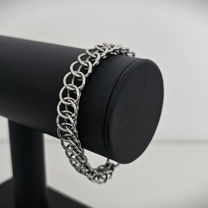 Puede incluir: Una pulsera de cota de malla plateada con un diseño de doble hebra. La pulsera se exhibe en un soporte cilíndrico negro para joyas sobre un fondo blanco. La pulsera está hecha de pequeños anillos de metal entrelazados.