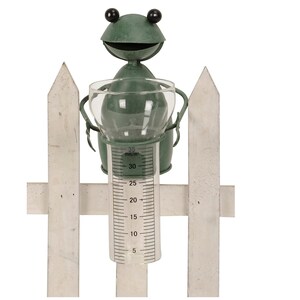 Könnte beinhalten: Ein skurriles Regenmessgerät in Form einer grünen Metallfroschfigur mit einem Messrohr aus klarem Glas. Der Frosch hält das Rohr, das mit Millimeter-Markierungen bis zu 35 mm versehen ist. Das Messgerät ist an einem weißen Zaun befestigt.