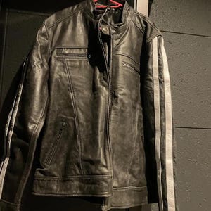 Peut inclure: Veste en cuir noir avec col montant et fermeture &eacute;clair sur le devant. La veste pr&eacute;sente deux bandes blanches sur chaque manche et plusieurs poches zipp&eacute;es. Le cuir a un aspect vieilli.