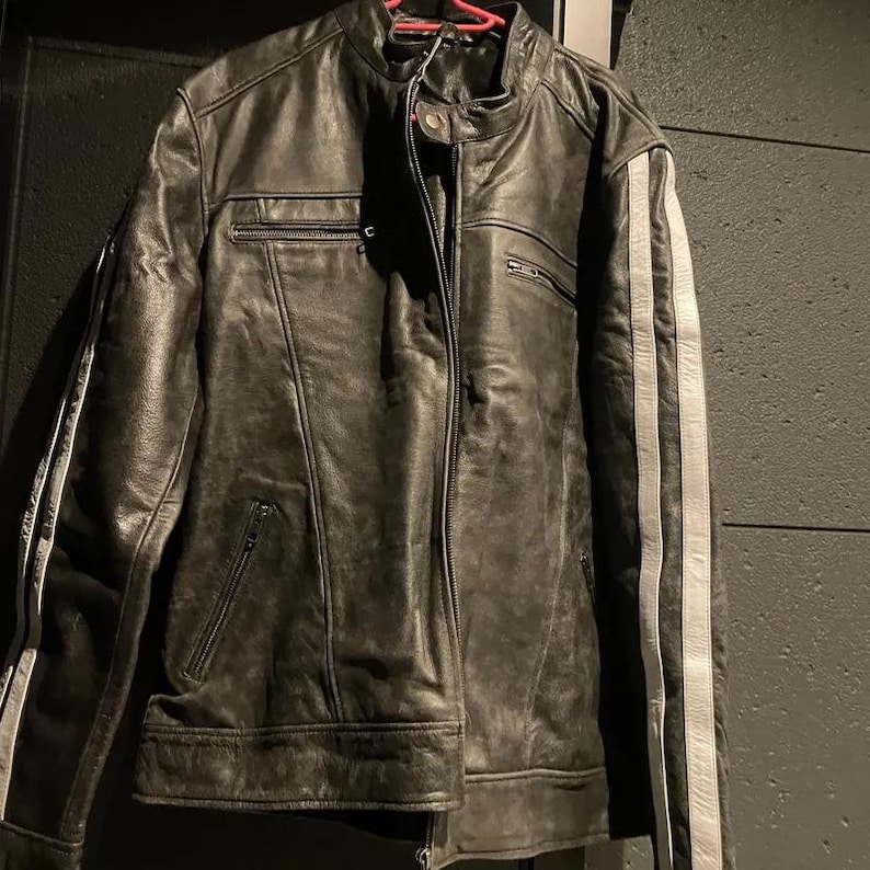 Peut inclure: Veste en cuir noir avec col montant et fermeture &eacute;clair sur le devant. La veste pr&eacute;sente deux bandes blanches sur chaque manche et plusieurs poches zipp&eacute;es. Le cuir a un aspect vieilli.