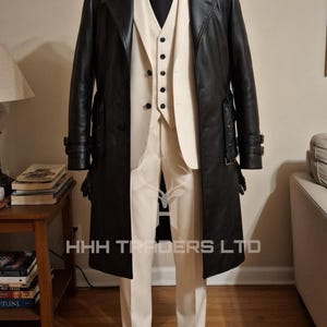 Puede incluir: Un abrigo largo de cuero negro, un traje color crema y una corbata negra se exhiben en un maniqu&iacute;. El traje incluye un chaleco y pantalones. El texto "HHH TRADERS LTD" es visible. Zapatos negros completan el conjunto.
