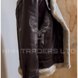 Pode incluir: Casaco de couro castanho com forro e acabamento em pele de carneiro creme. O casaco tem um estilo aviador cl&aacute;ssico com gola larga e fecho de correr. O texto "HHH TRADERS LTD" &eacute; vis&iacute;vel.