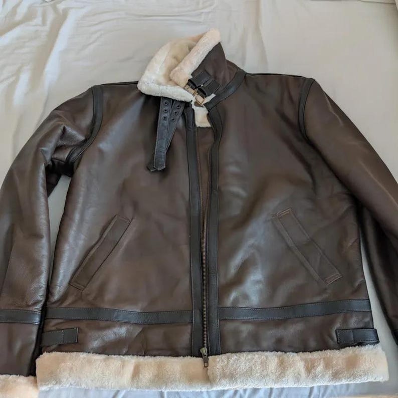 Pode incluir: Casaco de couro castanho com forro e gola de pele de ovelha creme. O casaco tem fecho de correr frontal, dois bolsos frontais e uma correia com fivela na gola. Estilo aviador cl&aacute;ssico.