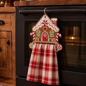 May include: A gingerbread house-shaped kitchen towel holder made of felt, featuring embroidered details like a red heart, candy canes, and peppermint swirls, with a red and white plaid fabric towel hanging from the base.