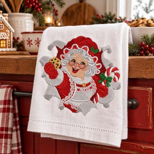 May include: A white kitchen towel featuring an embroidered design of Mrs. Claus wearing a red hat and coat, holding a chocolate chip cookie in one hand and a red and white striped candy cane in the other, surrounded by silver ribbon accents.