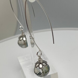 May include: Silver-tone dangle earrings featuring faceted, gray crystal teardrop-shaped beads. The beads are capped with a decorative silver filigree design. The earrings hang from curved silver wires, creating a delicate and elegant look.