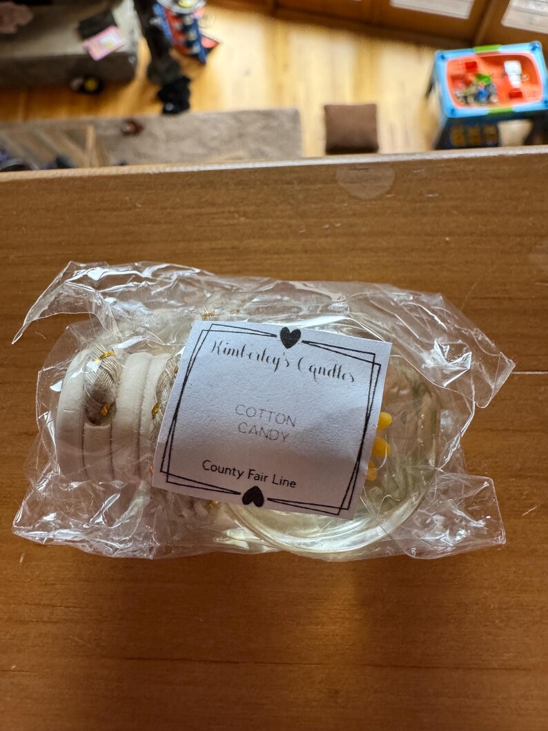 May include: A clear glass car air freshener bottle with a wooden cap and a white label that reads Kimberly's Candles, Cotton Candy, and County Fair Line, all enclosed in a transparent plastic bag with a hanging cord.
