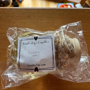 May include: A clear glass jar candle with a light-coloured braided rope handle and a wooden lid, sealed in a plastic bag. A white label features the text Kimberly's Candles, CARAMEL APPLE, and County Fair Line, decorated with two small black hearts.