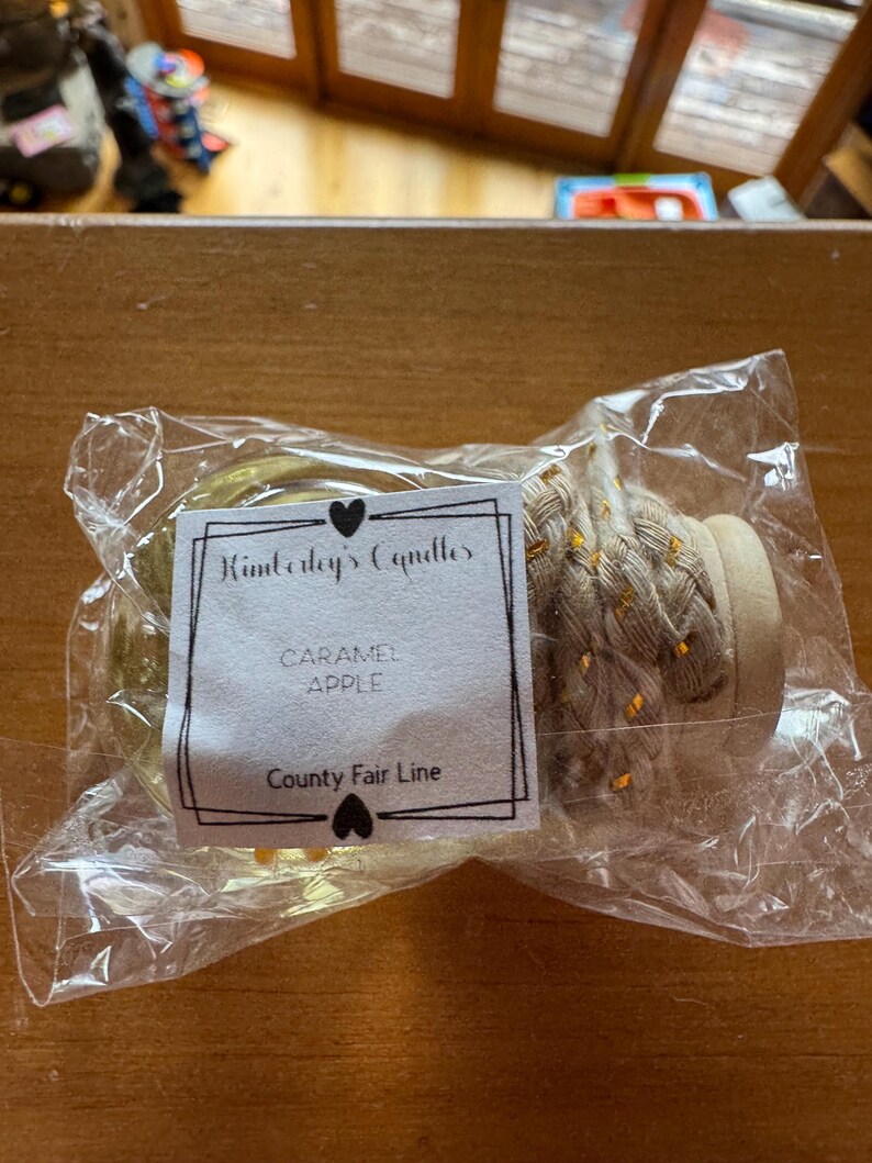 May include: A clear glass jar candle with a light-coloured braided rope handle and a wooden lid, sealed in a plastic bag. A white label features the text Kimberly's Candles, CARAMEL APPLE, and County Fair Line, decorated with two small black hearts.