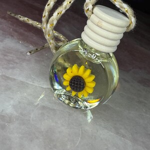 May include: A small, round, clear glass bottle filled with liquid, featuring a yellow sunflower charm with a dark brown centre inside. The bottle has a ribbed, light-coloured wooden cap and is suspended by a braided beige cord with gold metallic threads.
