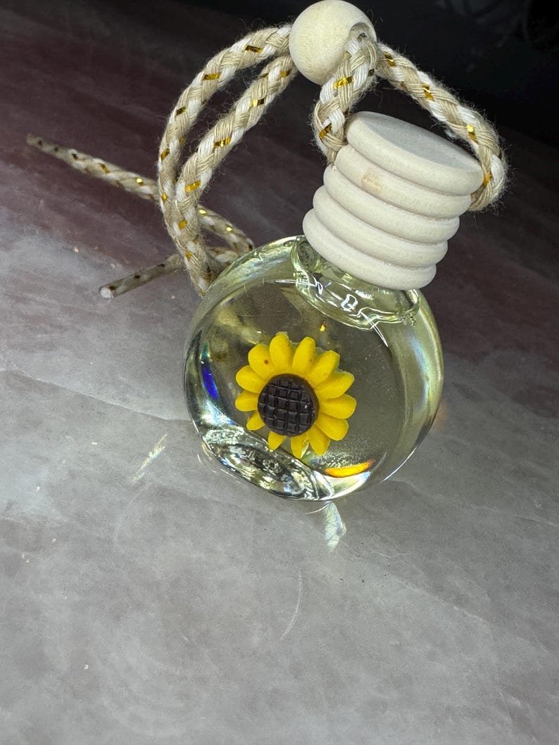 May include: A small, round, clear glass bottle filled with liquid, featuring a yellow sunflower charm with a dark brown centre inside. The bottle has a ribbed, light-coloured wooden cap and is suspended by a braided beige cord with gold metallic threads.