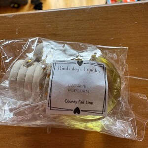 May include: A clear plastic-wrapped candle with a white label reading Kimberley's Candles, CARAMEL POPCORN, and County Fair Line. The candle features a light yellow wax base and a beige, textured, multi-layered top section with small gold accents.