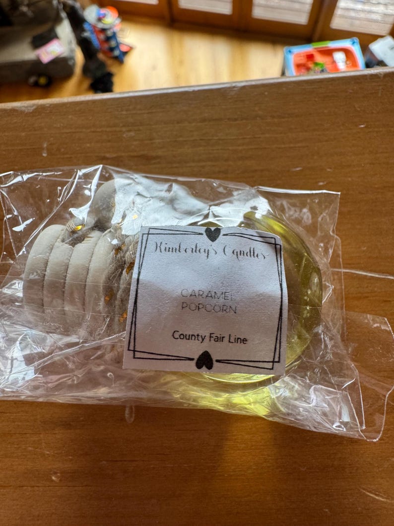 May include: A clear plastic-wrapped candle with a white label reading Kimberley's Candles, CARAMEL POPCORN, and County Fair Line. The candle features a light yellow wax base and a beige, textured, multi-layered top section with small gold accents.