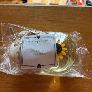 May include: A clear plastic-wrapped candle featuring a white label with the text Kimberly's Candles and APPLE BUTTER CARAMEL. The candle is shaped like a pumpkin with a braided texture and a small yellow sunflower accent on the side.