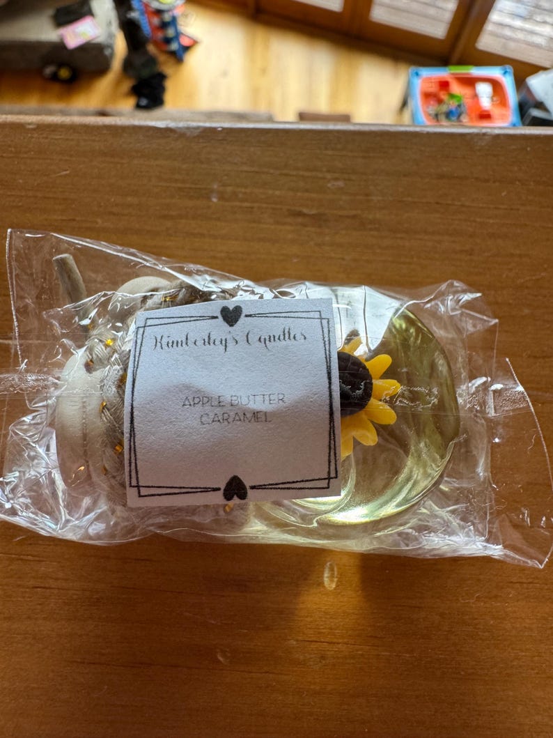 May include: A clear plastic-wrapped candle featuring a white label with the text Kimberly's Candles and APPLE BUTTER CARAMEL. The candle is shaped like a pumpkin with a braided texture and a small yellow sunflower accent on the side.