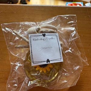 May include: A small glass car air freshener bottle filled with yellow liquid, featuring a yellow sunflower charm inside. It has a wooden cap with a hanging cord and a white label that reads Kimberly's Candles and CINNAMON, all in a clear plastic bag.