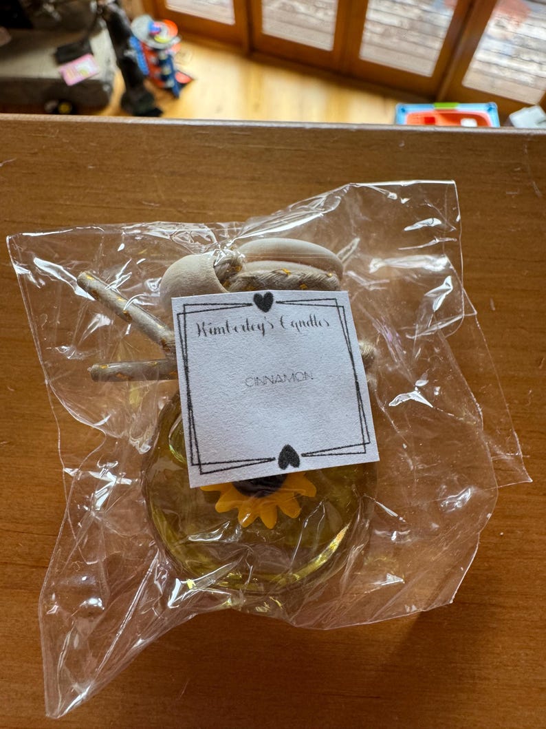 May include: A small glass car air freshener bottle filled with yellow liquid, featuring a yellow sunflower charm inside. It has a wooden cap with a hanging cord and a white label that reads Kimberly's Candles and CINNAMON, all in a clear plastic bag.