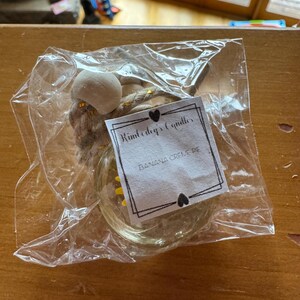 May include: A small glass jar candle with a thick rope hanger and a wooden bead, enclosed in a clear plastic bag. A white paper label is attached, featuring the text Kimberly's Candles and BANANA CREME PIE, framed by a black border with hearts.