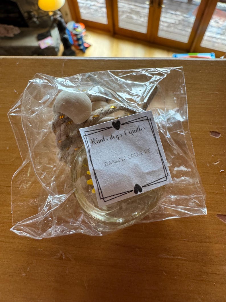 May include: A small glass jar candle with a thick rope hanger and a wooden bead, enclosed in a clear plastic bag. A white paper label is attached, featuring the text Kimberly's Candles and BANANA CREME PIE, framed by a black border with hearts.