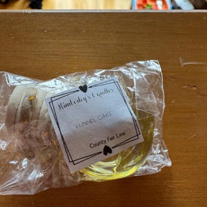 May include: A clear plastic-wrapped candle with a yellow wax base and a beige wick. The white rectangular label features black text reading Kimberley's Candles, FUNNEL CAKE, and County Fair Line, accented by two small black heart icons.