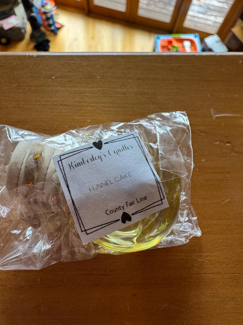 May include: A clear plastic-wrapped candle with a yellow wax base and a beige wick. The white rectangular label features black text reading Kimberley's Candles, FUNNEL CAKE, and County Fair Line, accented by two small black heart icons.