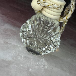 May include: A small, clear glass bottle with a faceted, shell-like texture. The bottle is topped with a light-coloured wooden cap and features a decorative braided cord with gold metallic threading for hanging, creating a distinct, reflective appearance.