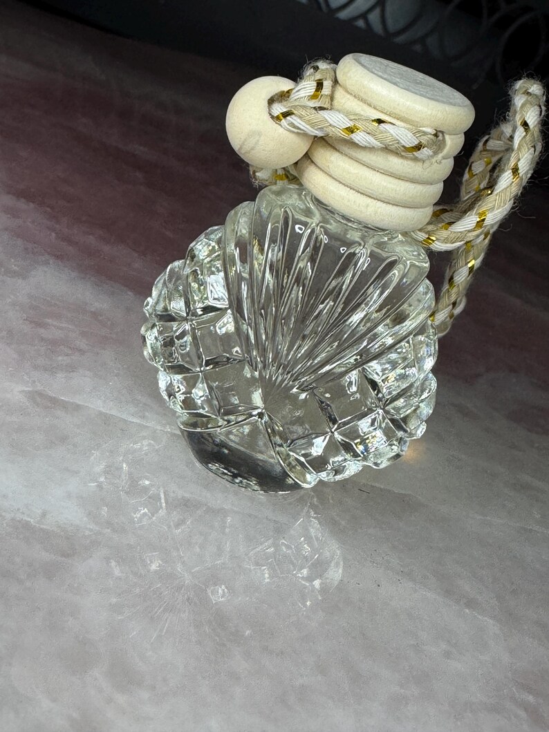 May include: A small, clear glass bottle with a faceted, shell-like texture. The bottle is topped with a light-coloured wooden cap and features a decorative braided cord with gold metallic threading for hanging, creating a distinct, reflective appearance.