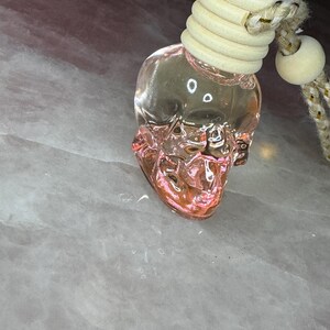 May include: A clear glass skull-shaped bottle filled with a pink liquid. The bottle features a ribbed light-coloured wooden cap and is attached to a thick, braided rope cord with a small wooden bead accent for hanging purposes.