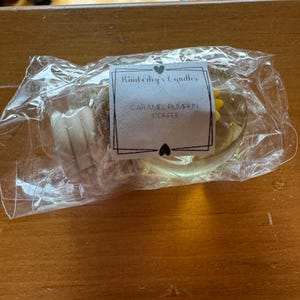 May include: A clear plastic-wrapped candle with a white label featuring the text Kimberley's Candles and Caramel Pumpkin Coffee. The candle is round, light-coloured, and includes a small yellow decorative element visible through the packaging.