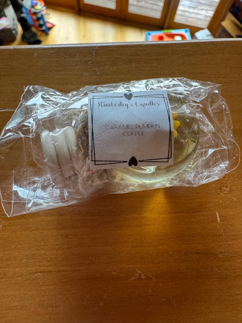 May include: A clear plastic-wrapped candle with a white label featuring the text Kimberley's Candles and Caramel Pumpkin Coffee. The candle is round, light-coloured, and includes a small yellow decorative element visible through the packaging.