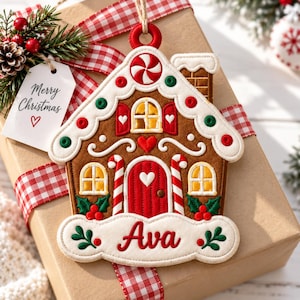 May include: A gingerbread house ornament featuring brown fabric, a red door, white icing details, candy cane accents, and green holly leaves. The base includes the name Ava embroidered in red. A tag attached to the ornament reads Merry Christmas.