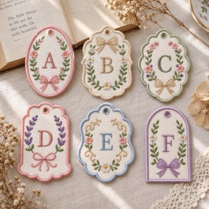 May include: Six embroidered fabric tags featuring letters A through F. Each tag has a unique shape, a central letter, and floral or bow motifs in pastel colors including pink, blue, green, and purple, with a small hole at the top for hanging.