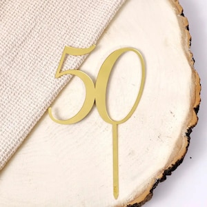 Op de afbeelding: Gouden metalen taarttopper in de vorm van het getal 50. De topper staat op een houten oppervlak.