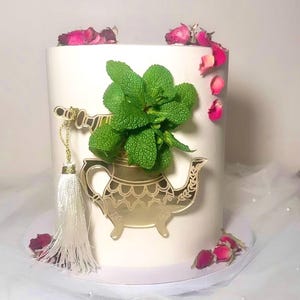 Könnte beinhalten: Eine weiße Torte, verziert mit einem goldenen Teekannen-Design und frischen Minzblättern. Rosa Rosenblätter sind um die Torte verstreut. Eine goldene Quaste hängt an dem Teekannen-Design.