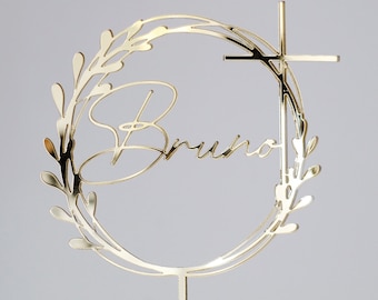 Pode incluir: Topper para bolo de metal dourado com um design de coroa circular e o nome "Bruno" escrito em itálico. Uma cruz está presa ao topo da coroa.